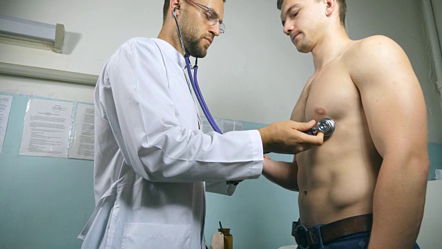 医生用听诊器检查年轻男性病人。医务工作者听着运动员的心跳。医生在医院的办公室里给一个人检查胸部。Close up Low angle view侧视图视频下载