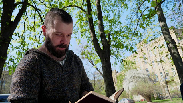 在一个阳光明媚的日子里，有胡子的男人坐在公园里读一本有趣的书视频素材