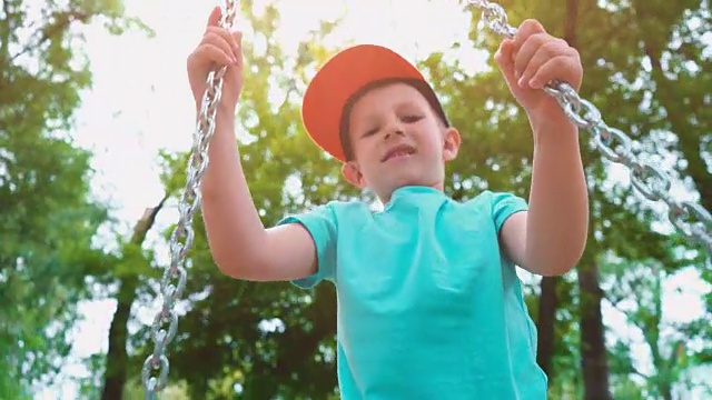 5岁的小男孩身穿蓝色t恤，头戴一顶帽子，带着钢链荡秋千，在绿树环绕的儿童秋千上玩得很开心视频素材