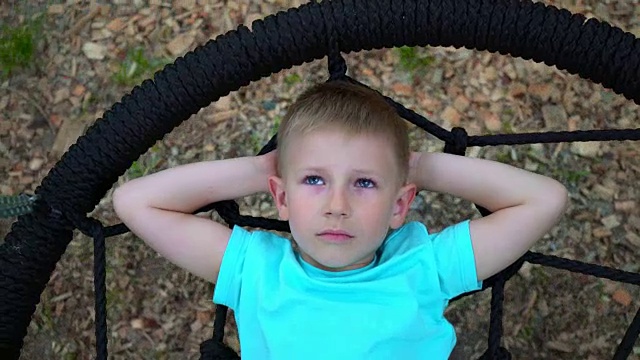 一个蓝眼睛的五岁男孩躺在秋千上，把手放在脑后，仰望着天空，梦想着，孩子正在休息，躺在秋千上视频素材