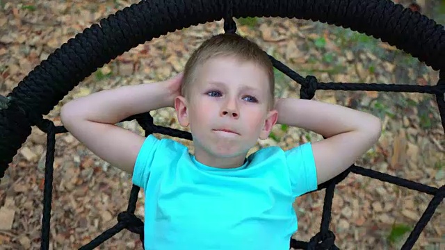 一个穿着蓝色t恤、蓝眼睛的五岁男孩躺在一个圆秋千上，把双手放在脑后，仰望着天空，梦想着，这个孩子正在休息，躺在秋千上视频下载