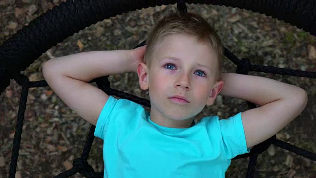 一个蓝眼睛的五岁男孩躺在秋千上，把手放在脑后，仰望着天空，梦想着，孩子正在休息，躺在秋千上视频素材