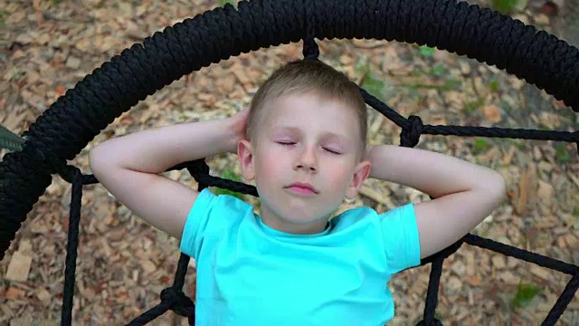 一个穿着蓝色t恤、蓝眼睛的5岁男孩闭着眼睛躺在一个圆形秋千上做梦。一个孩子在想一个愉快的盘旋在地面上的秋千时笑了视频素材
