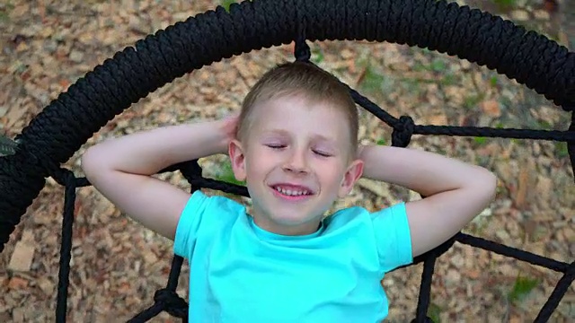 一个穿着蓝色t恤、蓝眼睛的5岁男孩闭着眼睛躺在一个圆形秋千上做梦。一个孩子在想一个愉快的盘旋在地面上的秋千时笑了视频下载