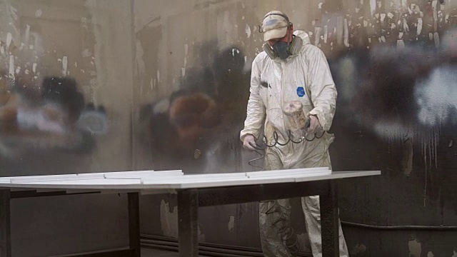 戴着防护面具和工作服的成熟男子在小制造厂为家具的细节上色视频素材