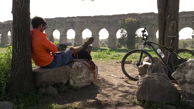 骑自行车的人坐在一棵树下的岩石上，在古罗马的渡槽前用他的笔记本电脑进行视频通话。年轻有吸引力的运动员与橙色运动装和背包parco degli acququedotti在罗马视频下载