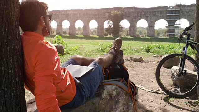 骑自行车的人坐在树下的一块岩石上，用他的笔记本电脑听音乐，在古罗马的渡槽前喝水。年轻有吸引力的运动员与橙色运动服耳机和背包parco degli acququedotti在罗马视频下载