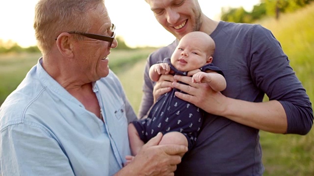 Multi-generation家庭。祖父亲吻新生孙子的脚。三个男人在河边的自然中休息。丰富多彩的日落视频下载