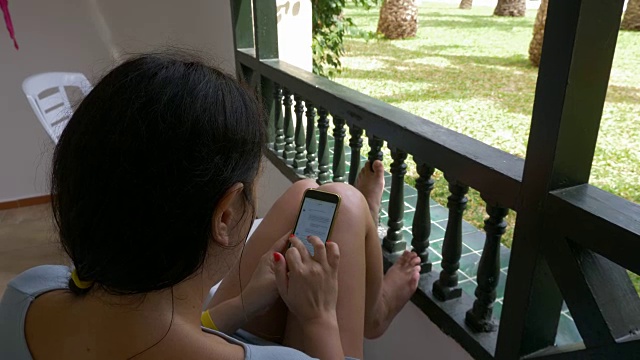 年轻女子坐在户外阳台上使用手机。女人在手机上发短信。肩视图高角度视频下载