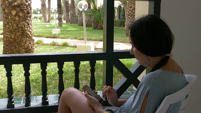 成年女性夏天在室外阳台休息时使用智能手机。现代女性在手机上看旅游博客视频下载
