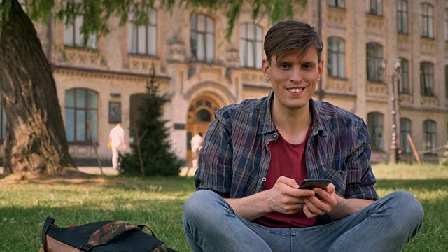 年轻帅气的男子坐在公园的草地上，轻拍智能手机，对着镜头走来走去，放松的概念，交流的概念，建立在背景上视频下载