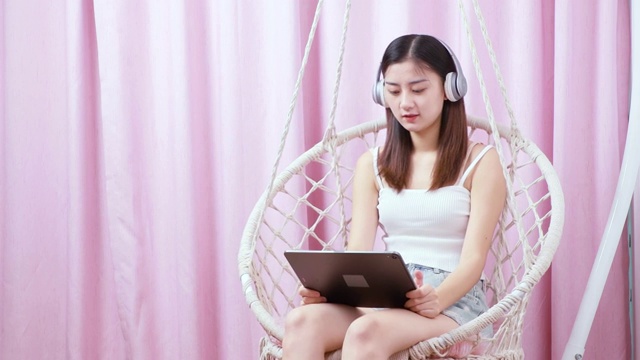 可爱的中国女孩，坐在吊椅上带着耳机用着平板电脑视频素材