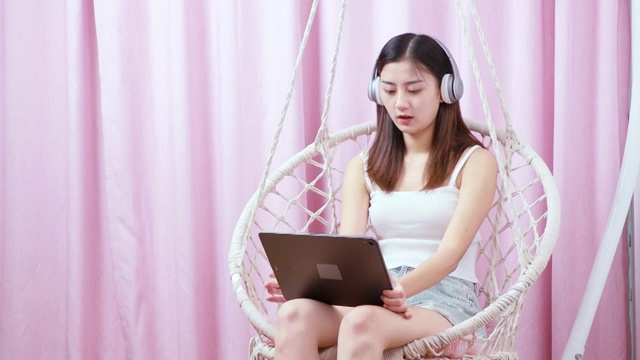 可爱的中国女孩，坐在吊椅上带着耳机看平板电脑视频素材