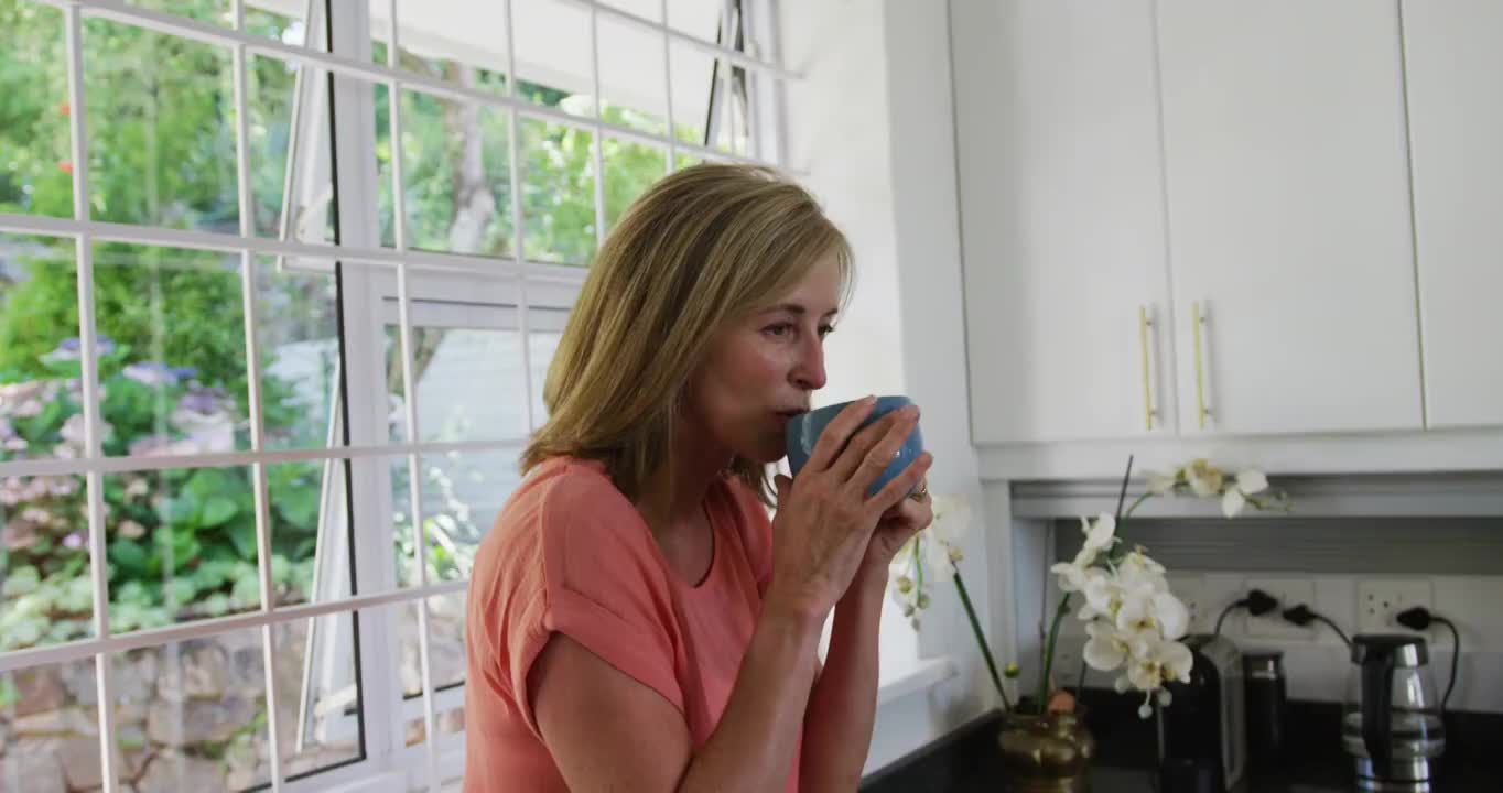 一个白人妇女站在家里的厨房里喝着咖啡视频素材