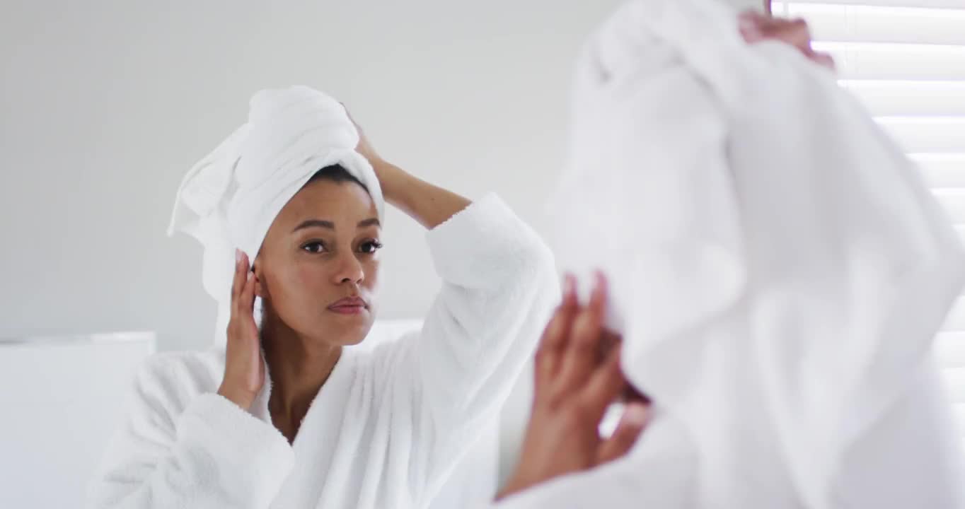 非裔美国妇女在浴衣触摸她的头在浴室看着镜子视频素材