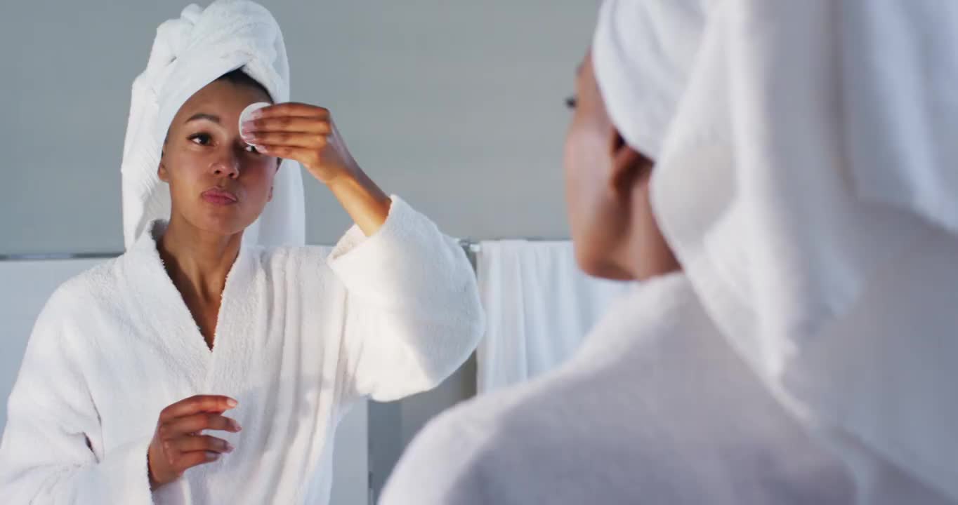 非裔美国妇女在浴衣清洁她的脸与棉垫在浴室看着镜子视频素材