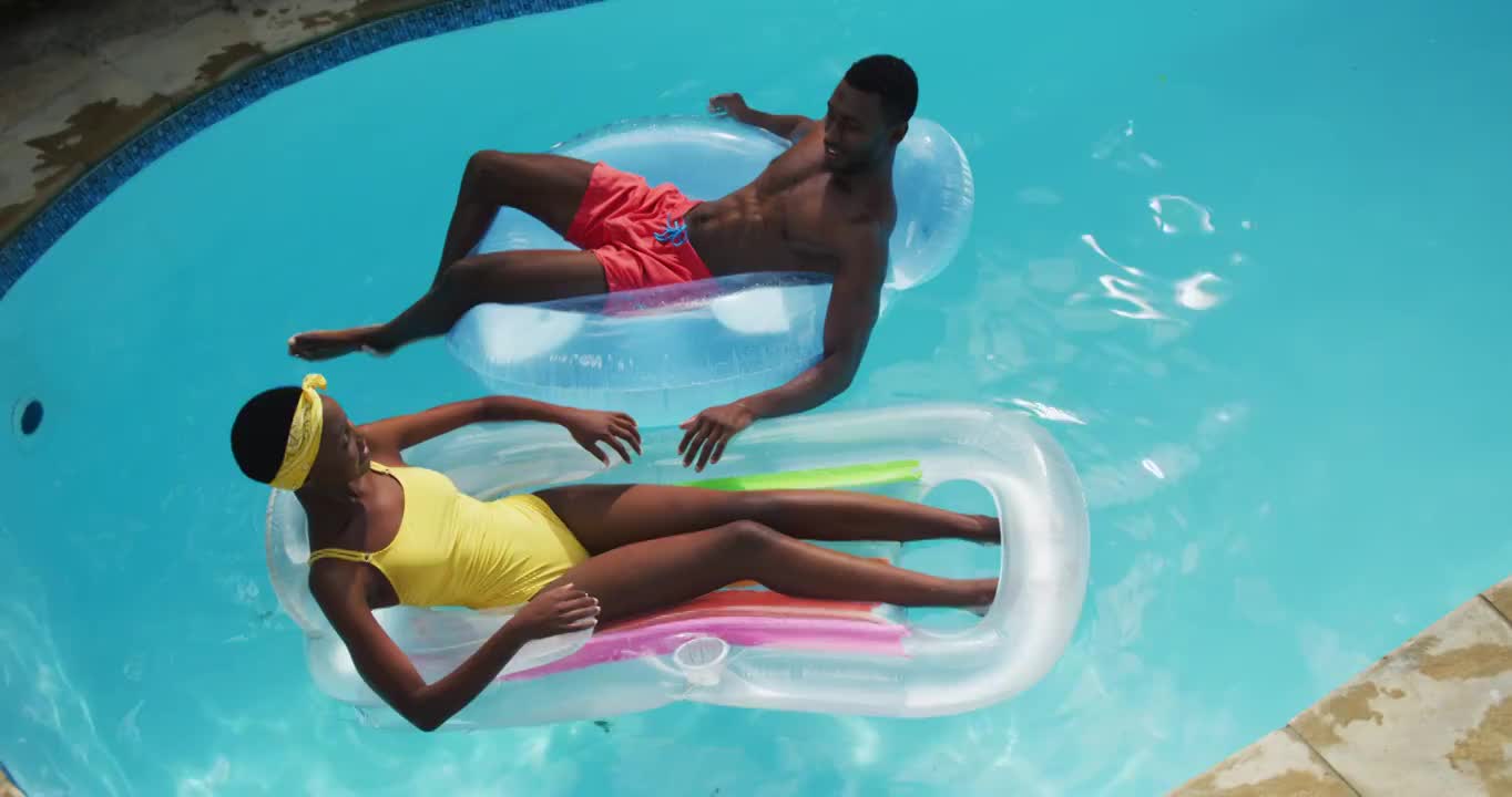 快乐的非裔美国夫妇躺在游泳池里的充气玩具上，微笑着互相溅起水花视频素材