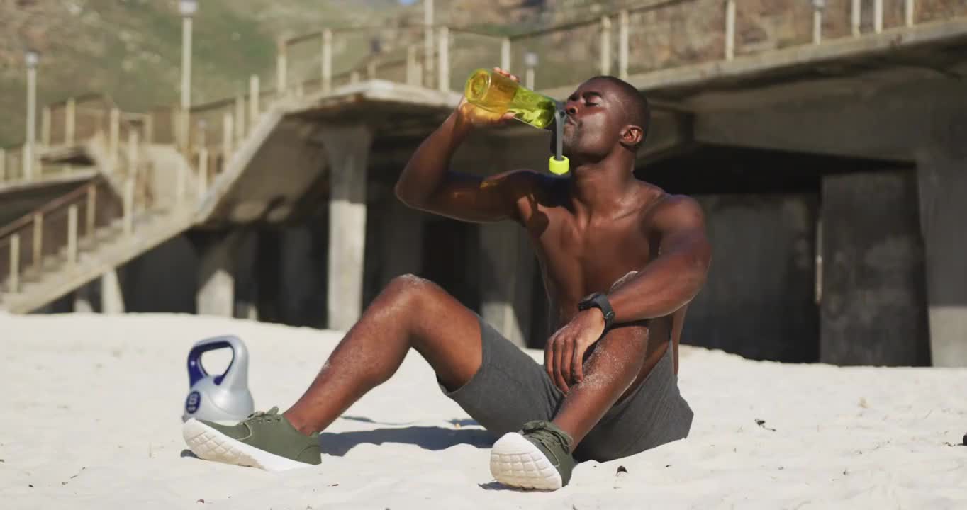 疲惫的非洲裔美国人坐在沙滩上，喝着瓶子里的水，在户外运动中休息视频下载