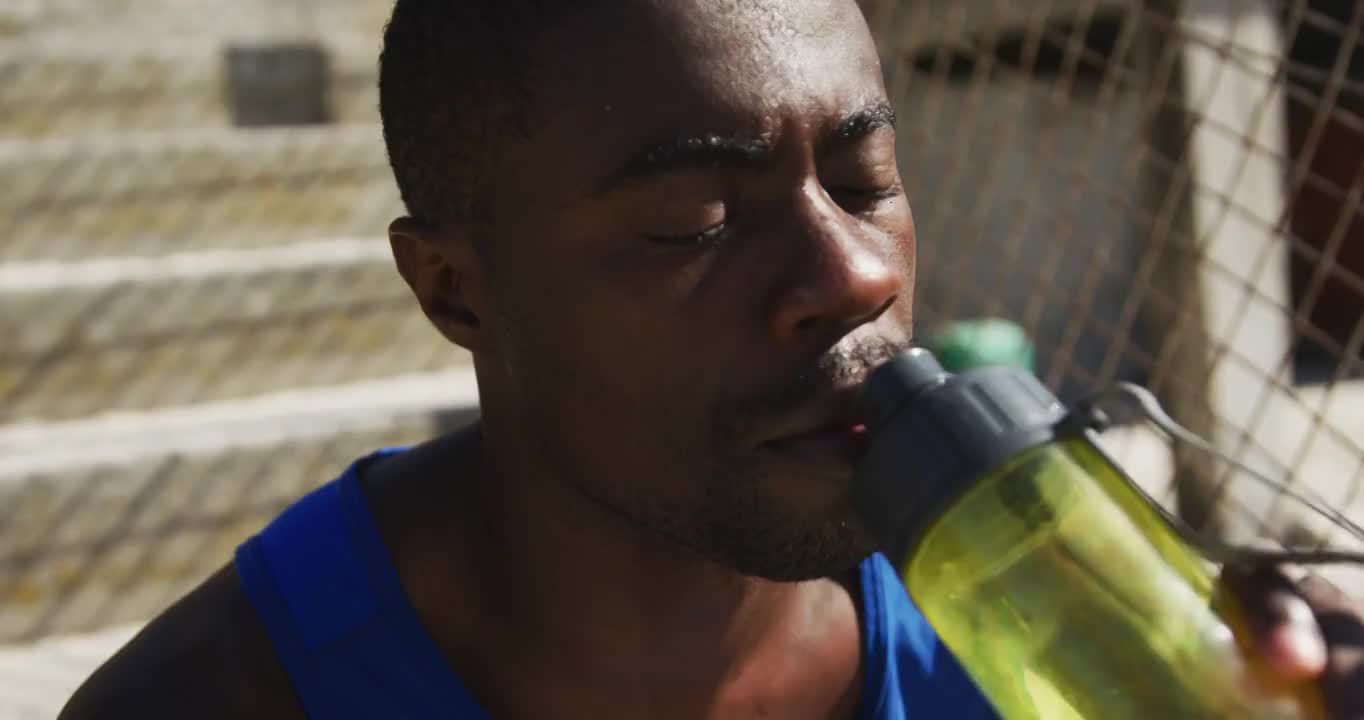 疲惫的非洲裔美国人坐在那里，喝着水壶里的水，在户外运动中休息视频素材