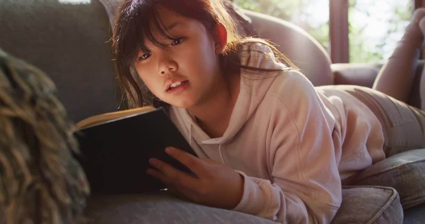 亚洲女孩微笑着在家里躺在沙发上看书视频下载