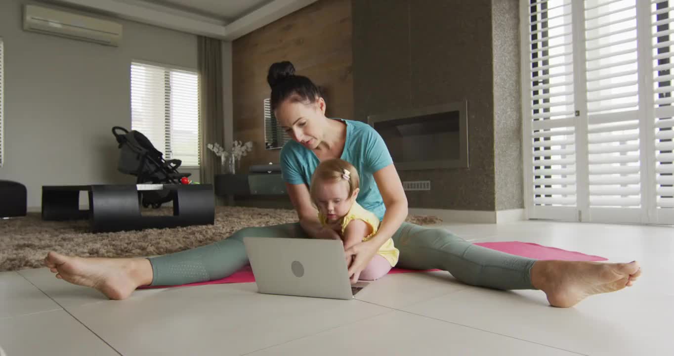 一位白人母亲抱着她的孩子在瑜伽垫上练习瑜伽，坐在家里的笔记本电脑前视频下载