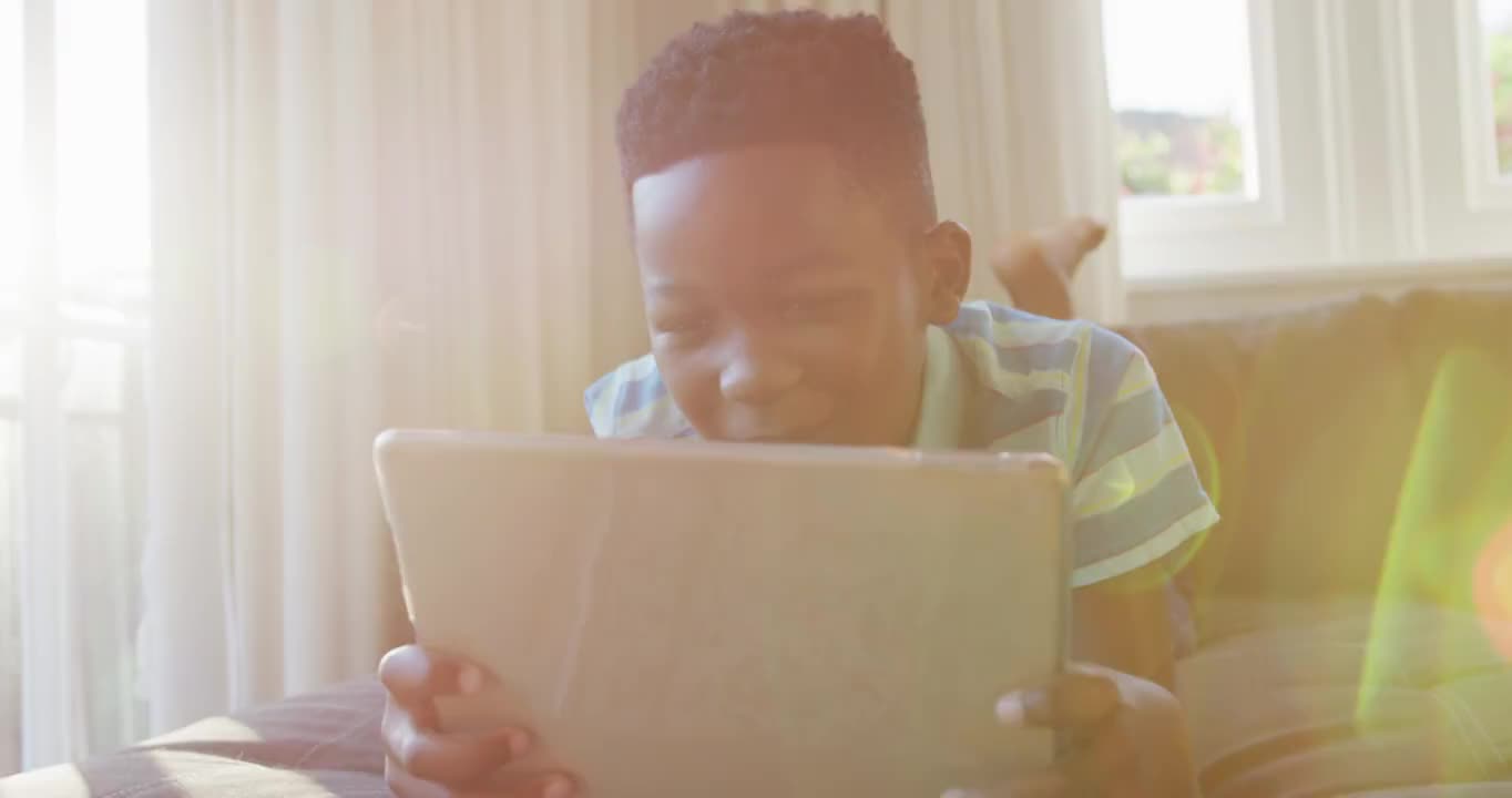 快乐的非洲裔美国男孩躺在沙发上使用平板电脑和微笑视频下载