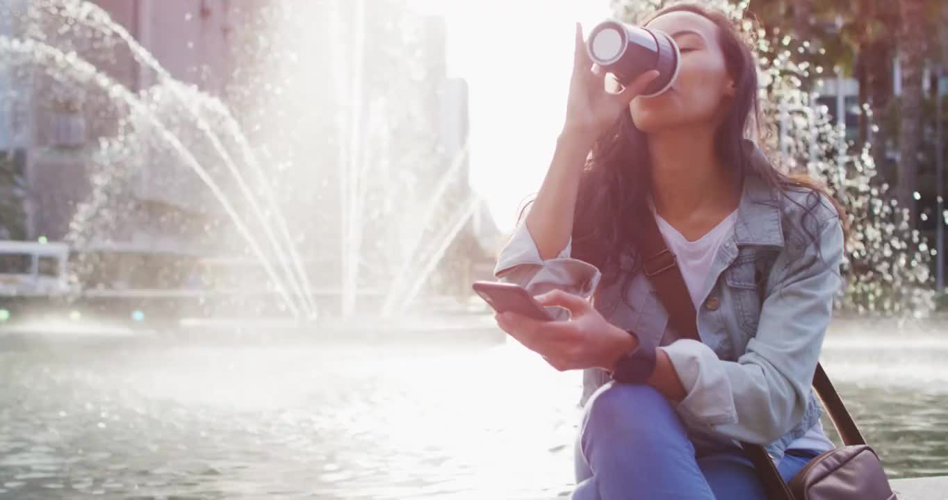 亚洲女性坐在喷泉旁边用智能手机喝外卖咖啡视频素材