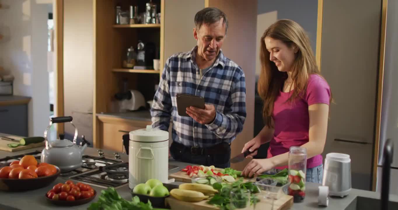 面带微笑的白人父亲和十几岁的女儿在厨房用平板电脑准备保健饮料视频素材