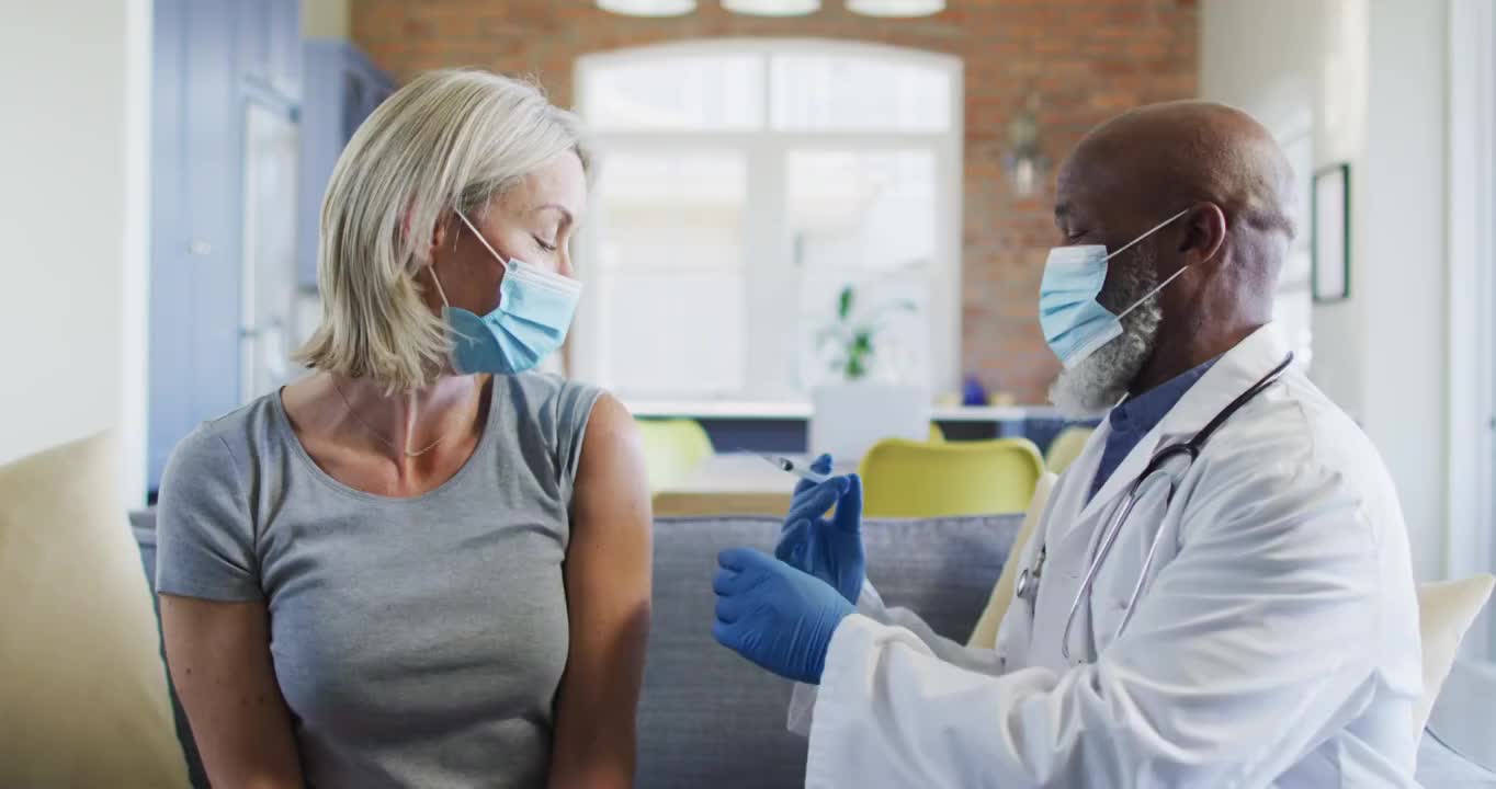 快乐的高级多样化的女人和医生戴着口罩坐在客厅的沙发上，接种疫苗视频下载
