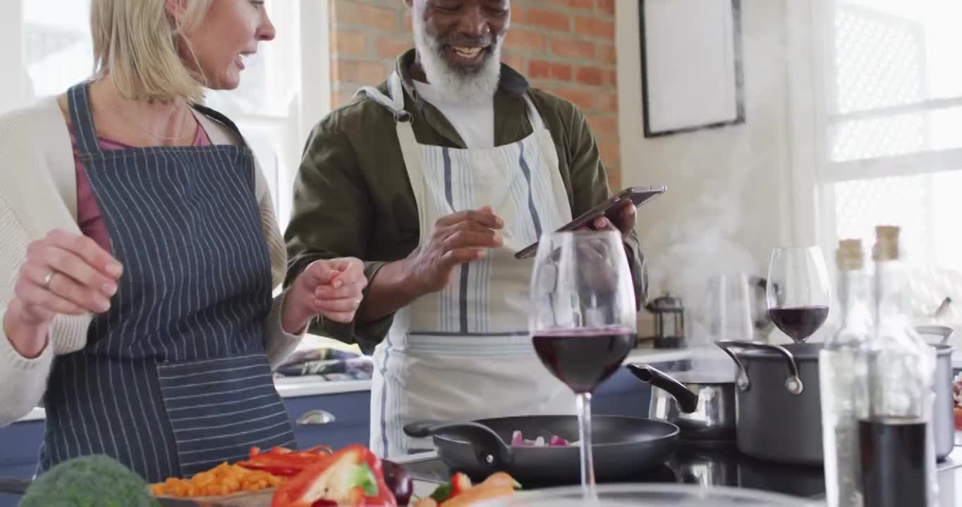 混合种族的老年夫妇穿着围裙，在家里的厨房里用数码平板电脑做饭视频素材