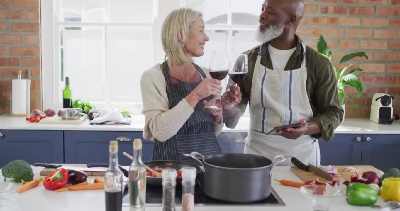 穿着围裙的混合种族老年夫妇在家里的厨房里喝酒做饭视频下载