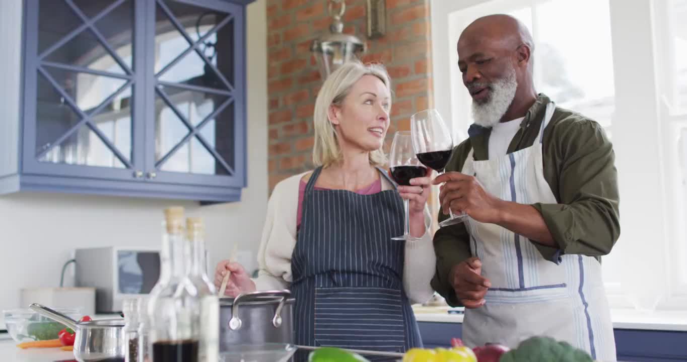 穿着围裙的混合种族老年夫妇在家里的厨房里喝酒做饭视频素材