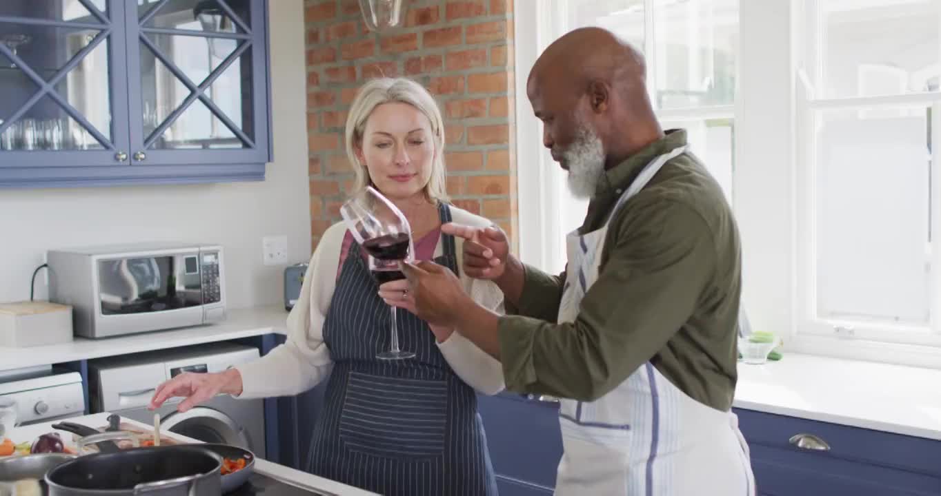 穿着围裙的混合种族老年夫妇在家里的厨房里用酒杯做饭视频素材