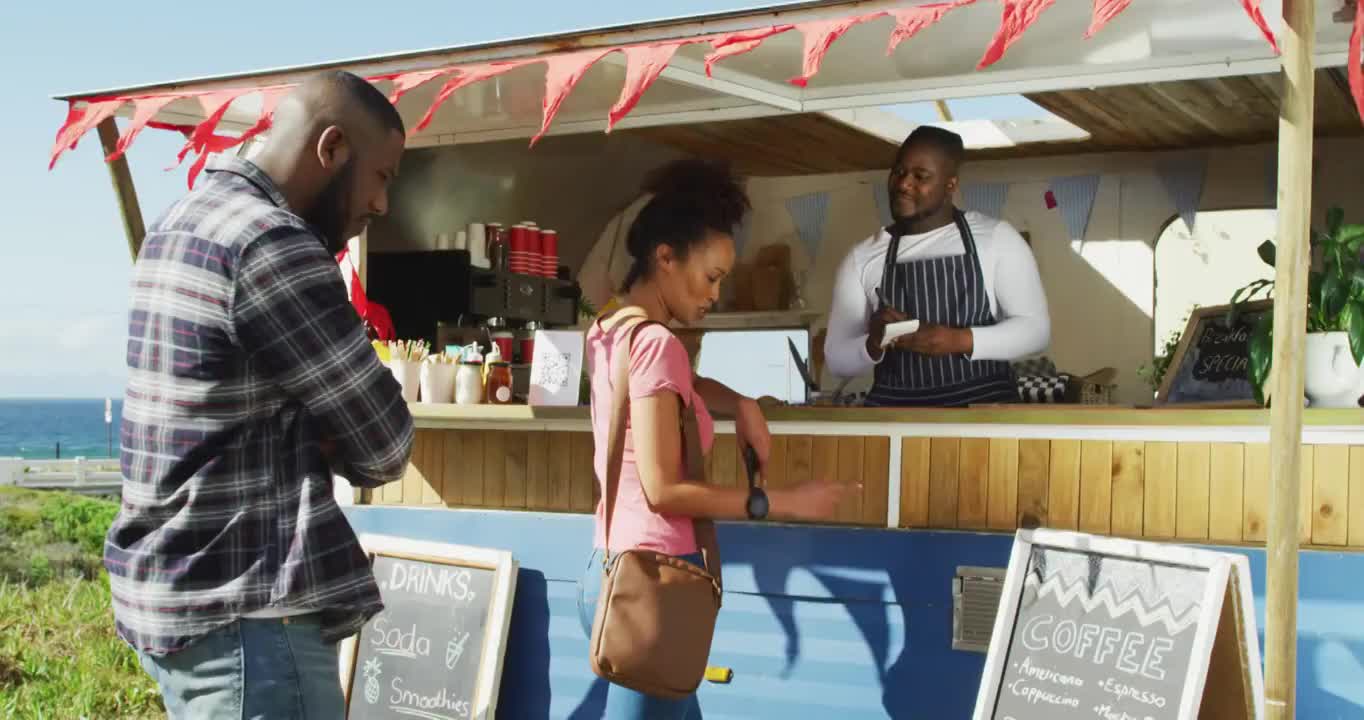 面带微笑的非洲裔美国男性餐车老板在接受女顾客点餐，男顾客在排队等候视频下载