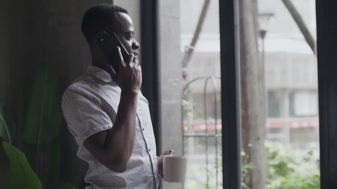 一位非洲黑人商人站在窗前喝咖啡，用手机交谈的侧视图视频素材