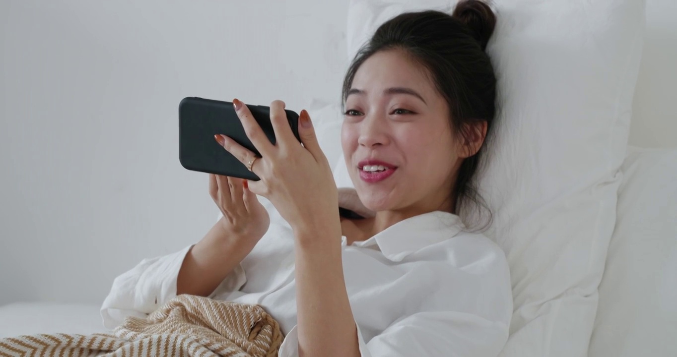 穿着白衬衫的年轻快乐的亚洲女孩躺在床上打视频电话和朋友聊天，视频素材