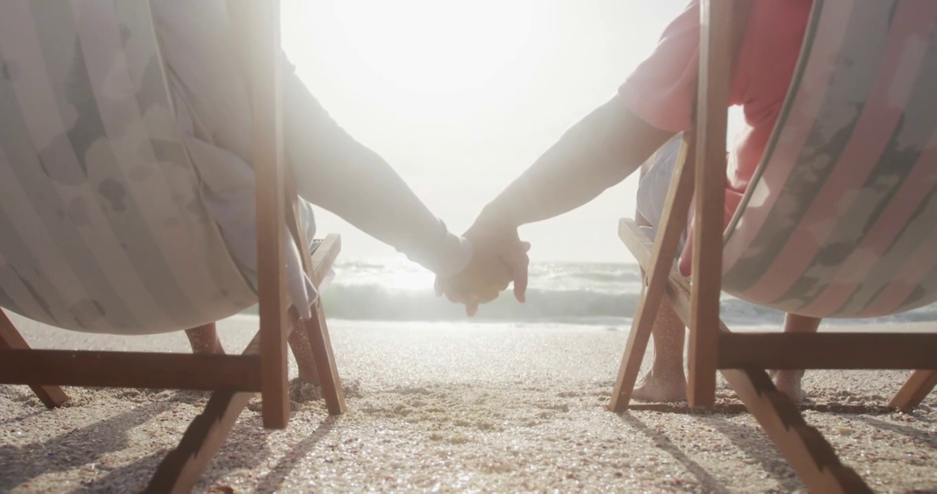 夕阳西下，西班牙老年夫妇在沙滩上的日光浴床上放松，手牵手的背影视频下载