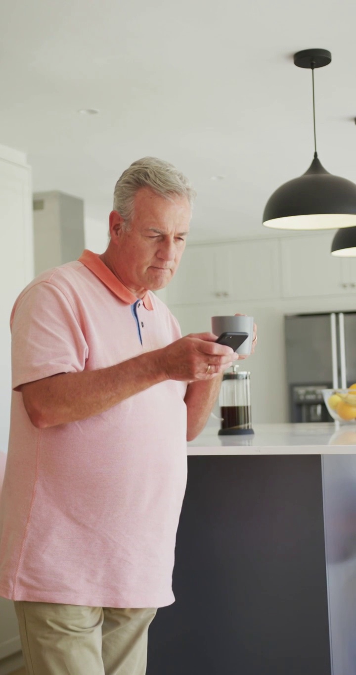 一名白人老人在厨房里一边喝咖啡一边用智能手机的垂直镜头视频素材