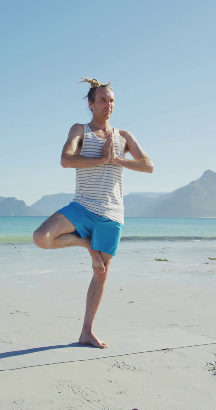 放松的白人男子与脏辫练习瑜伽站在海滩垂直视频视频下载