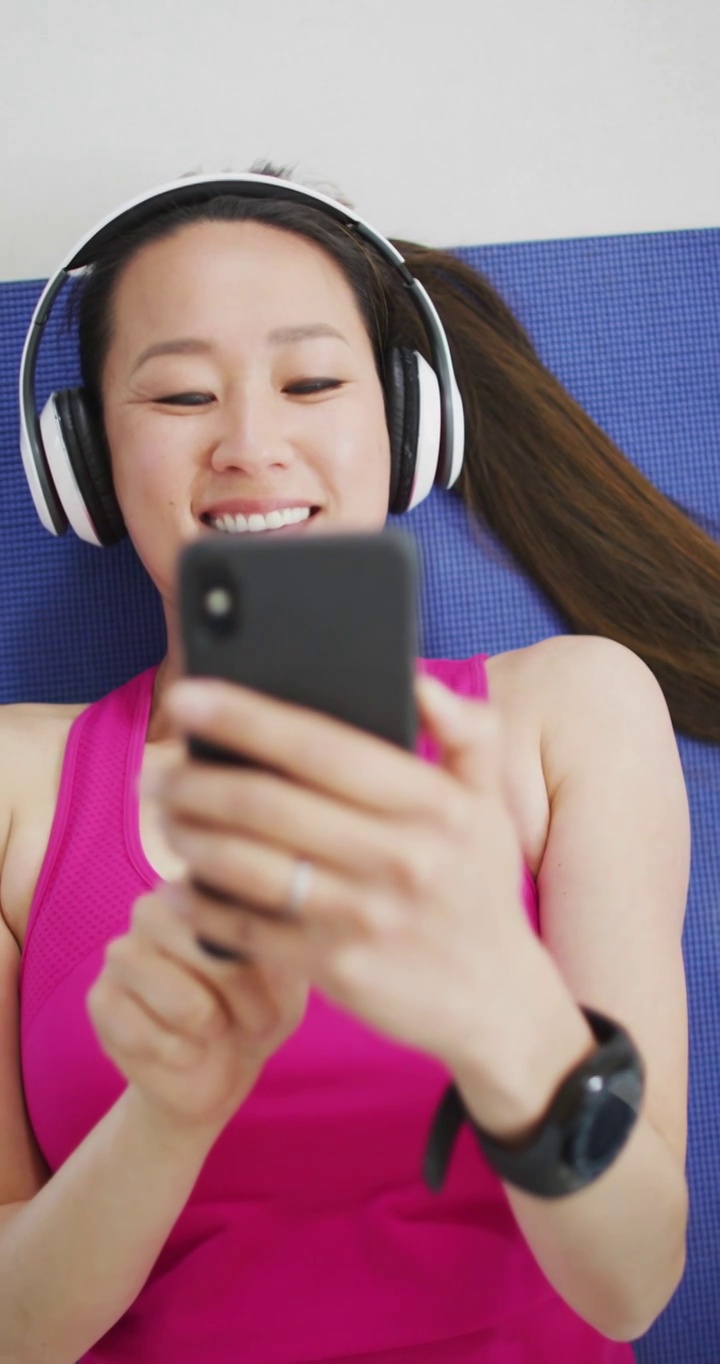 垂直视频微笑的亚洲女人放松戴耳机和使用智能手机视频下载