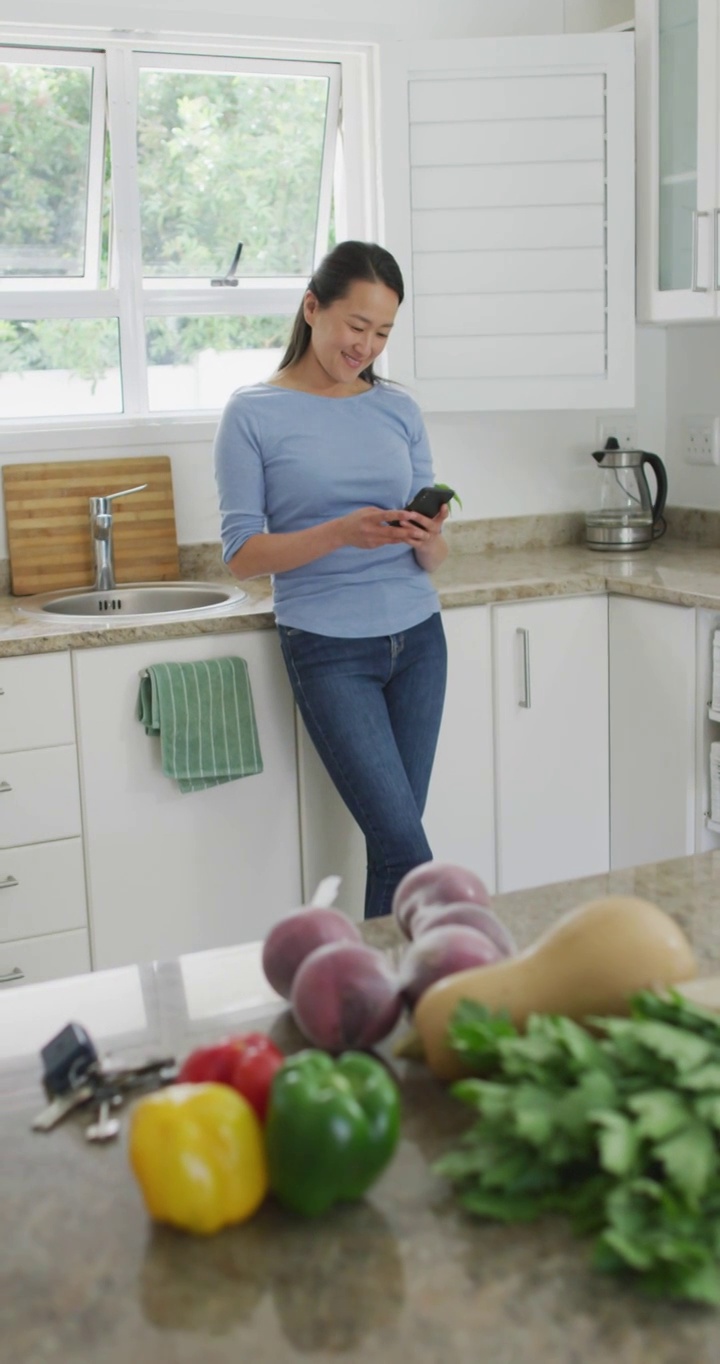 垂直视频微笑的亚洲妇女站在厨房使用智能手机视频下载