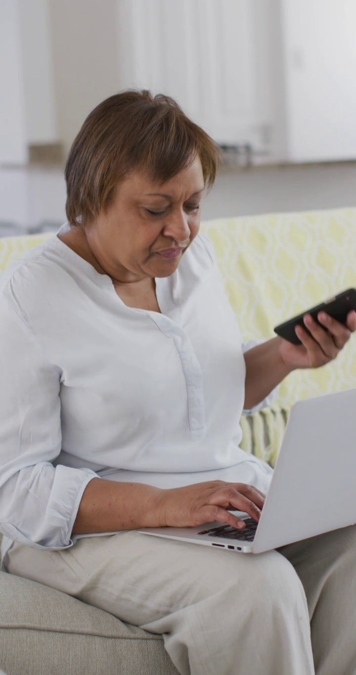 垂直视频微笑老年非洲裔美国妇女使用智能手机和笔记本电脑坐在家里视频下载