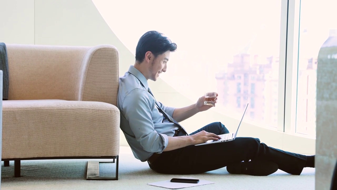 商务男子坐在酒店房间地上使用笔记本电脑工作、视频通话干杯庆祝成功视频下载