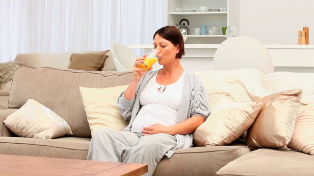 深色头发的孕妇坐在沙发上喝着橙汁视频下载