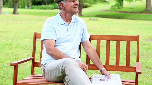 在一个阳光明媚的日子里，一个退休男子在公园长椅上拿着报纸放松视频下载