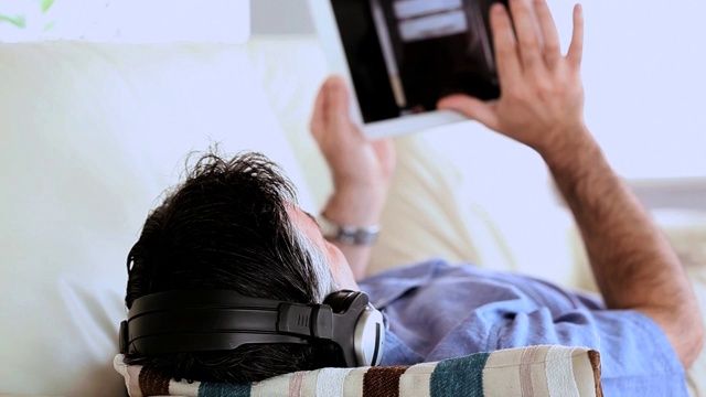 一个成熟的男人躺在沙发上听着音乐，在客厅里用着平板电脑视频素材