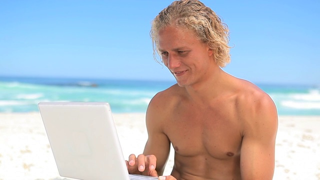 微笑的金发男人拿着笔记本电脑在海滩上视频下载