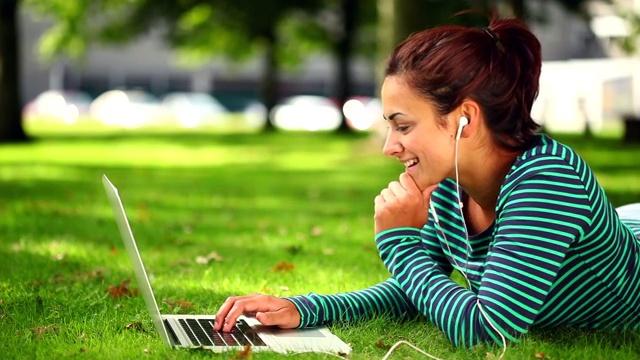 漂亮的学生躺在草地上用笔记本电脑在大学里听音乐视频素材