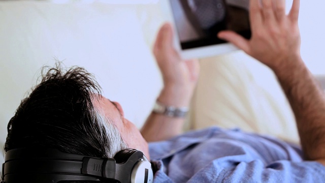 一个成熟的男人躺在沙发上听音乐，在客厅里用平板电脑视频素材