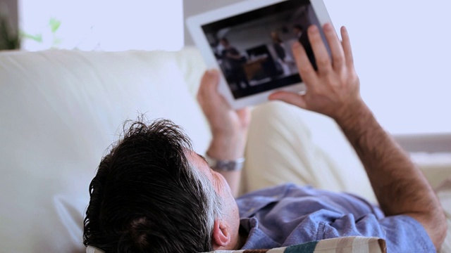 一个成熟的男人躺在沙发上，在客厅里用着平板电脑视频素材